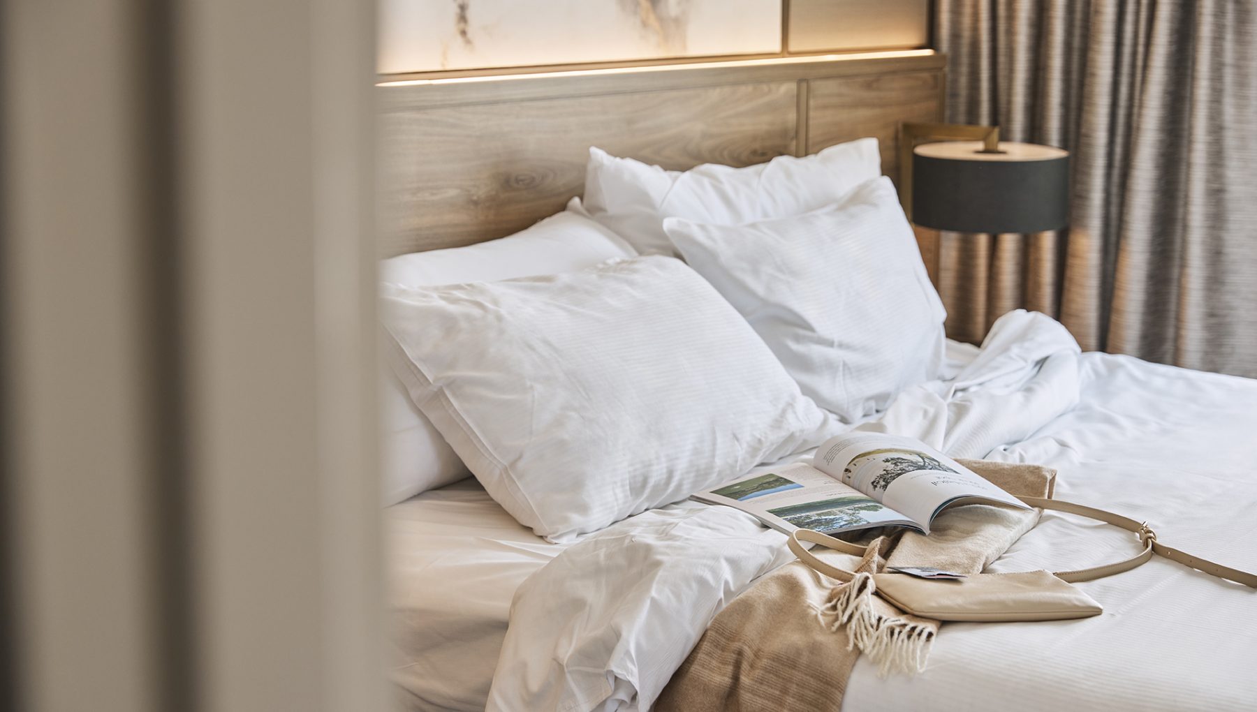 Unmade hotel bed with white pillows, an open magazine, a beige bag and throw, beside a dark bedside lamp.
