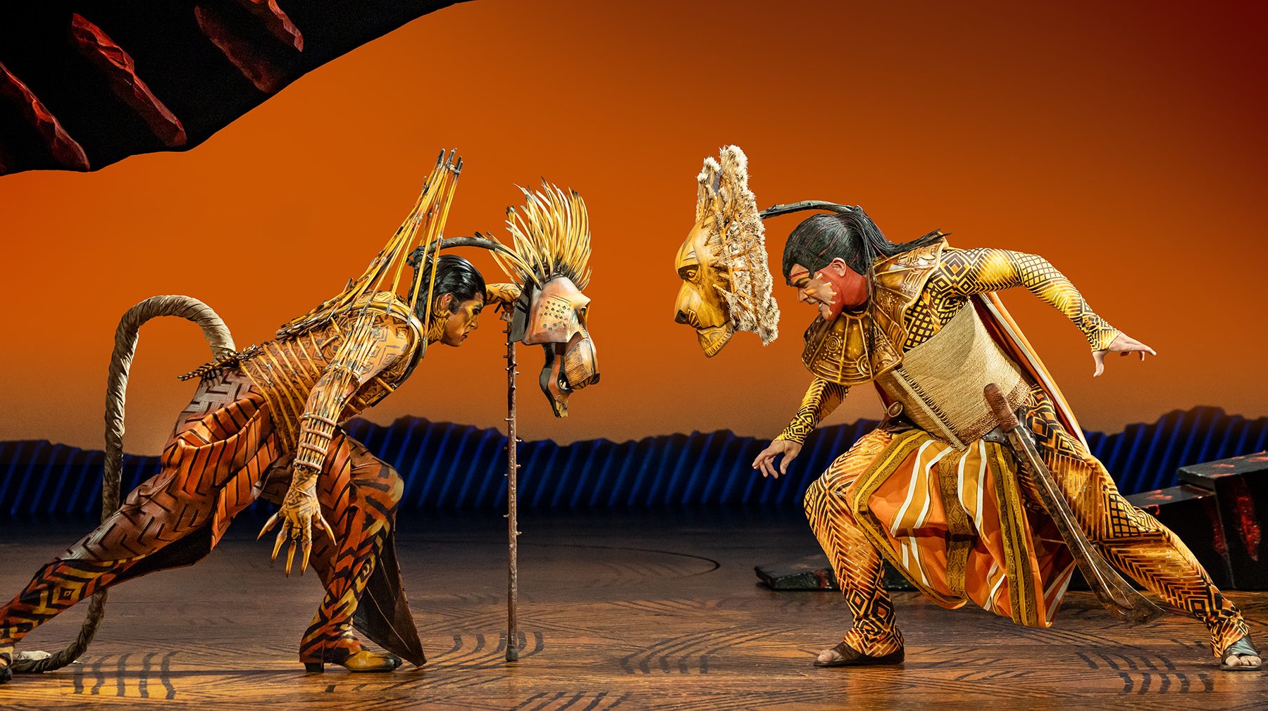 Two performers in ornate orange-and-gold costumes crouch facing each other onstage, with feathered masks displayed between them.