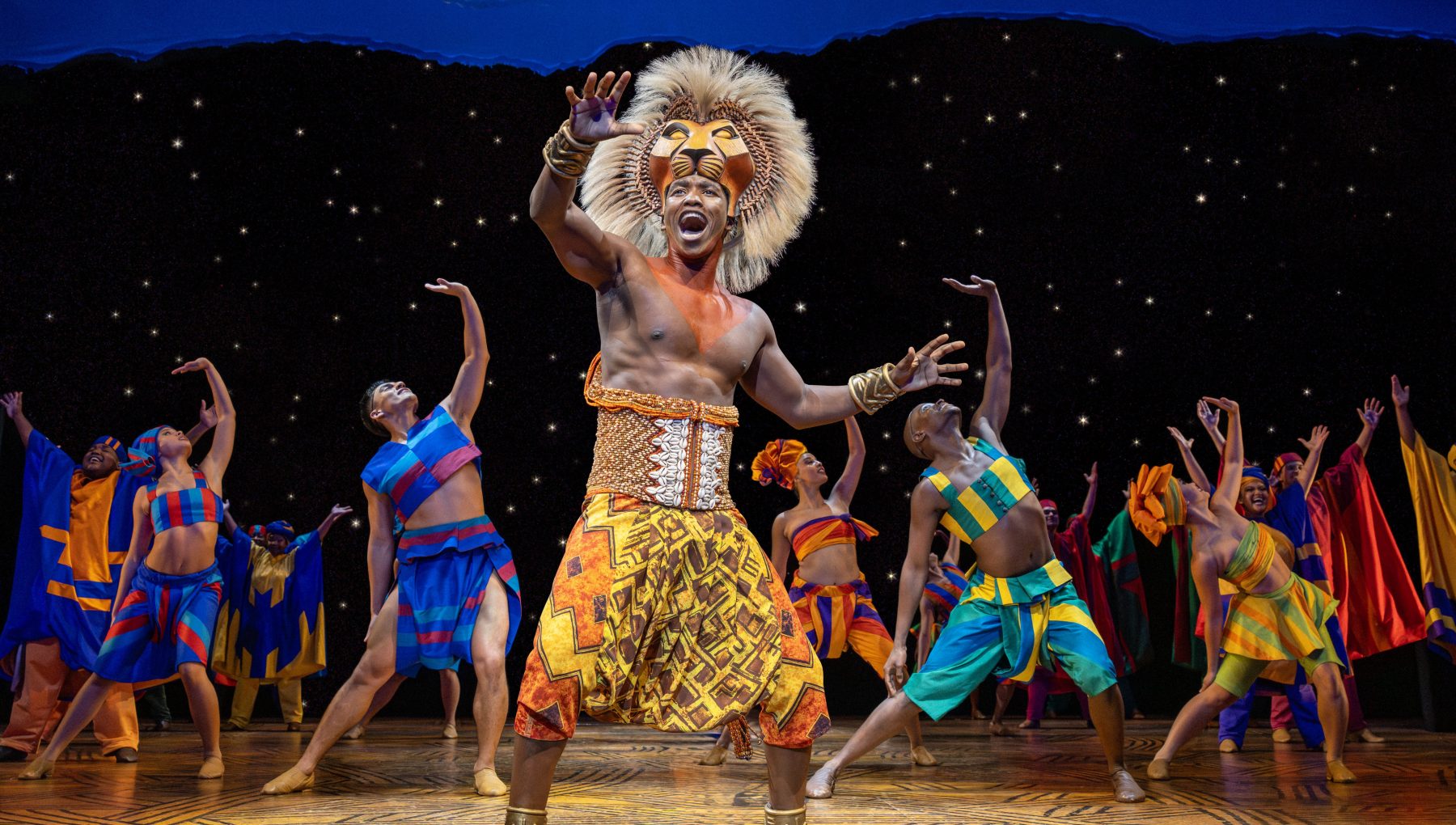 A performer in a feathered lion headdress and beaded costume raises an arm as dancers in colorful outfits perform on a starry stage.