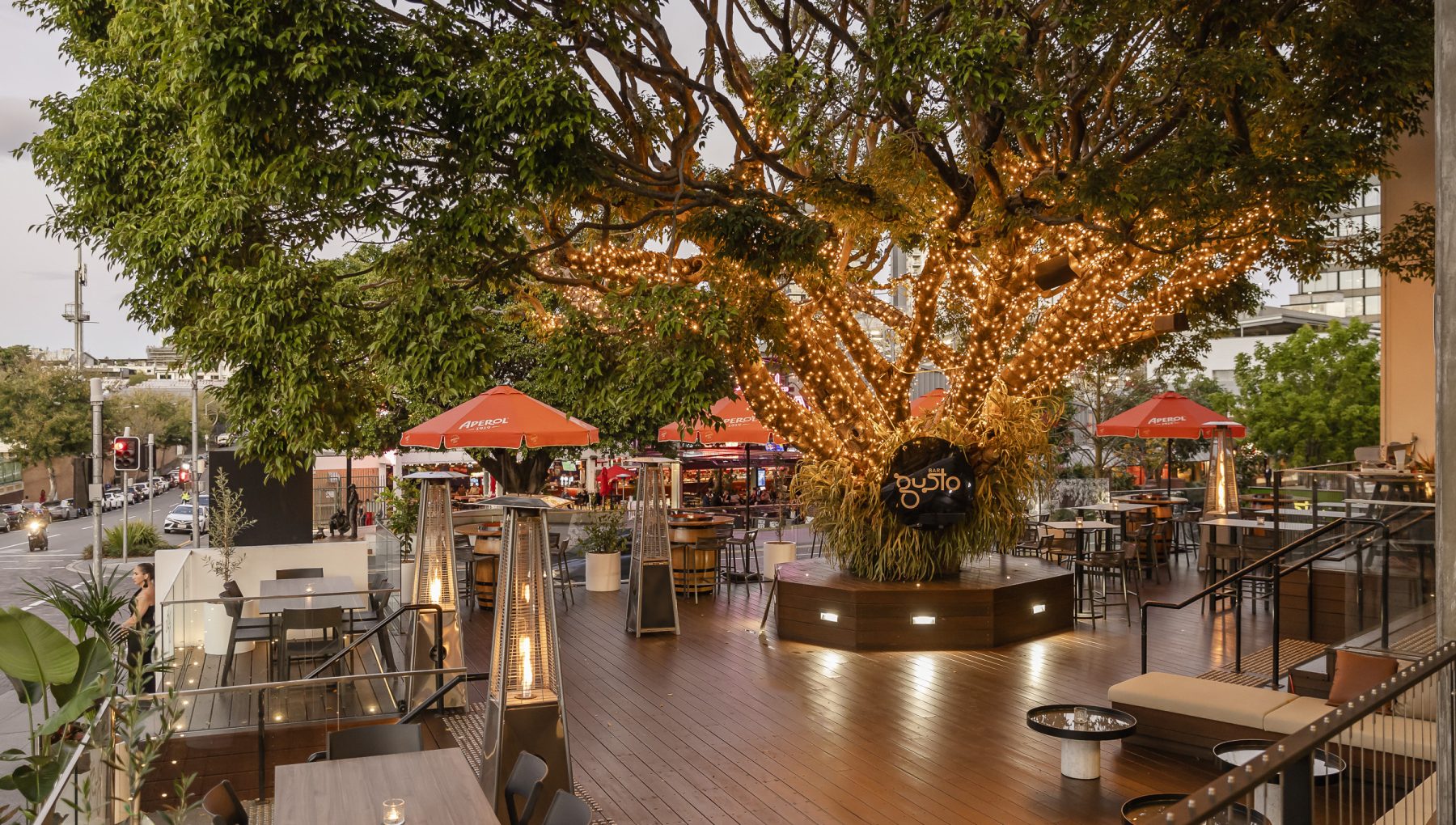 A vibrant outdoor dining area features a large tree adorned with string lights, surrounded by tables and chairs under orange umbrellas.