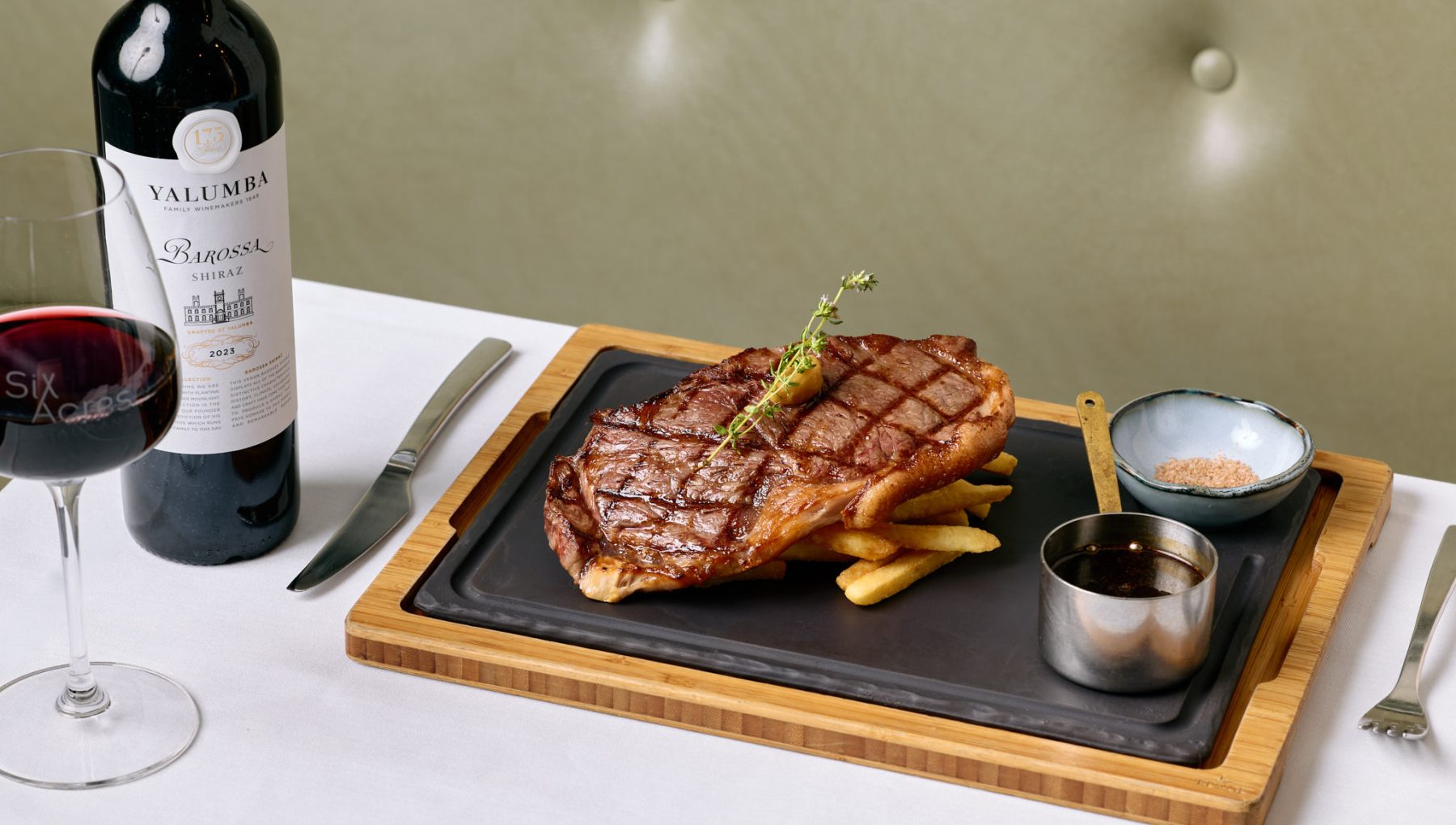Grilled steak with fries, dipping sauces, a glass of red wine, and a wine bottle on a dining table.
