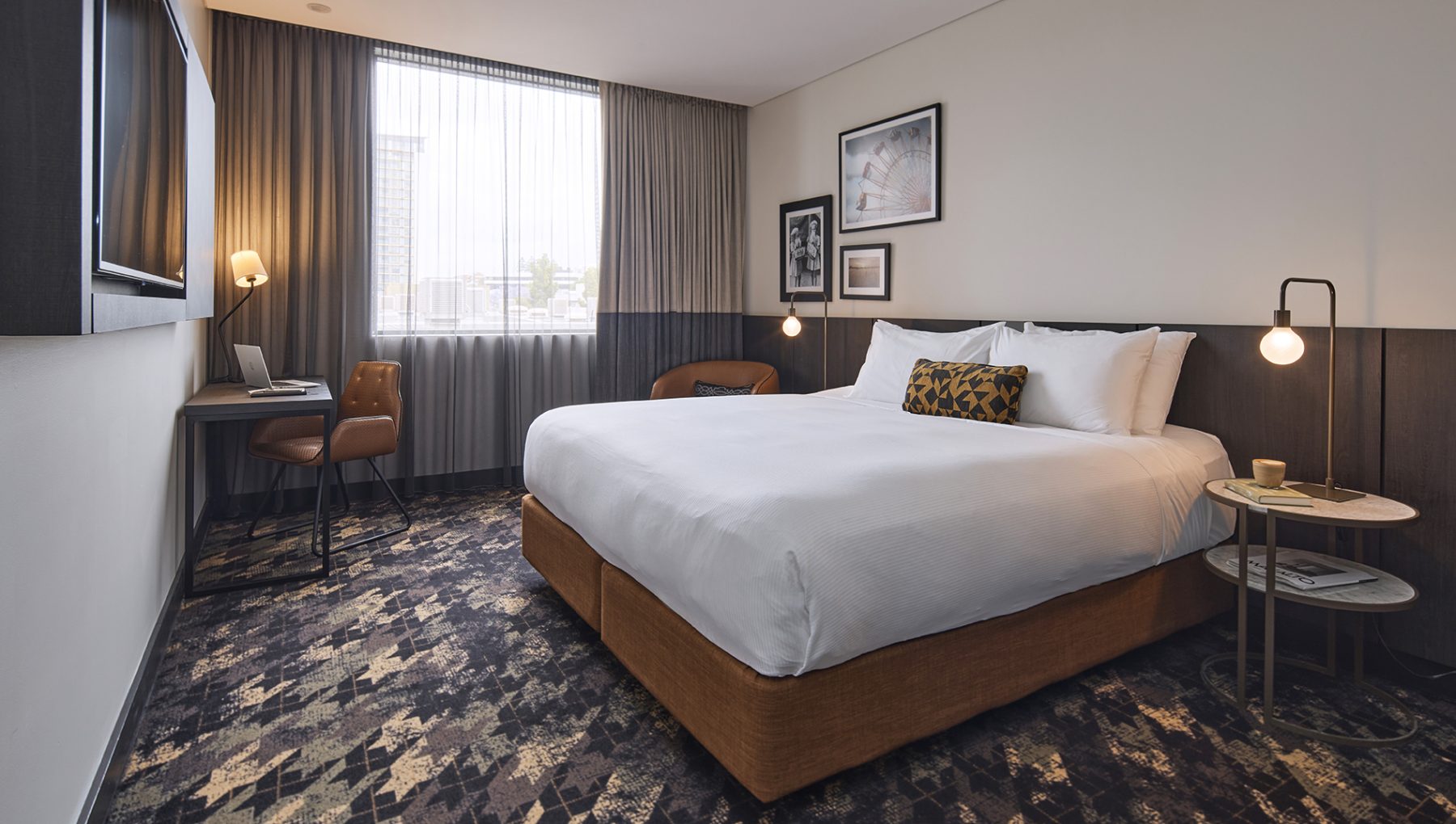Modern hotel room with a large white bed, bedside tables and lamps, desk and chair, framed art, and a curtained window.