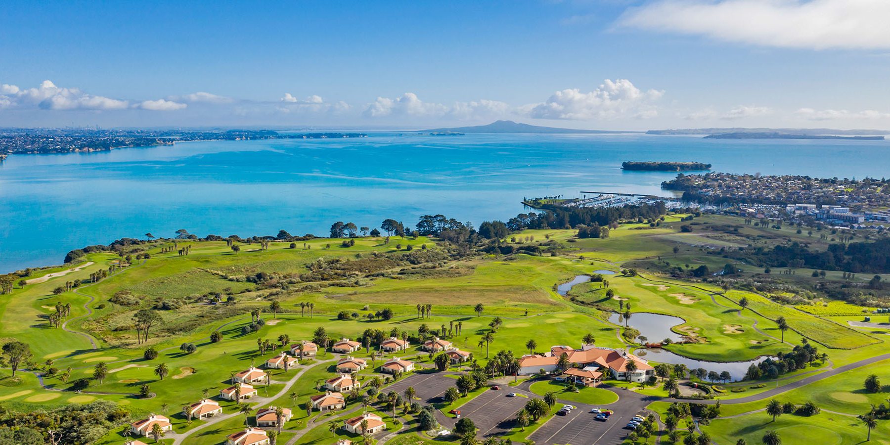 Scenic aerial view of a golf course with green lawns, palm trees, and a blue coastal landscape under a clear sky.