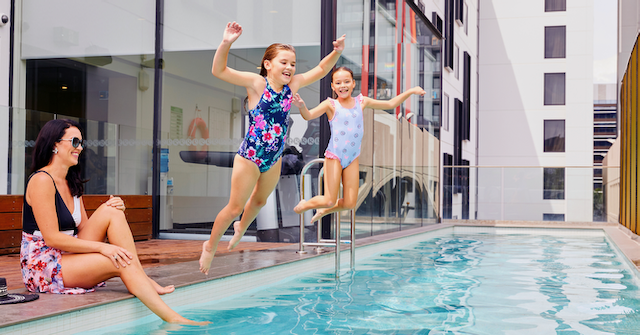 Two girls in swimsuits joyfully leap into a swimming pool while a woman relaxes on the edge.