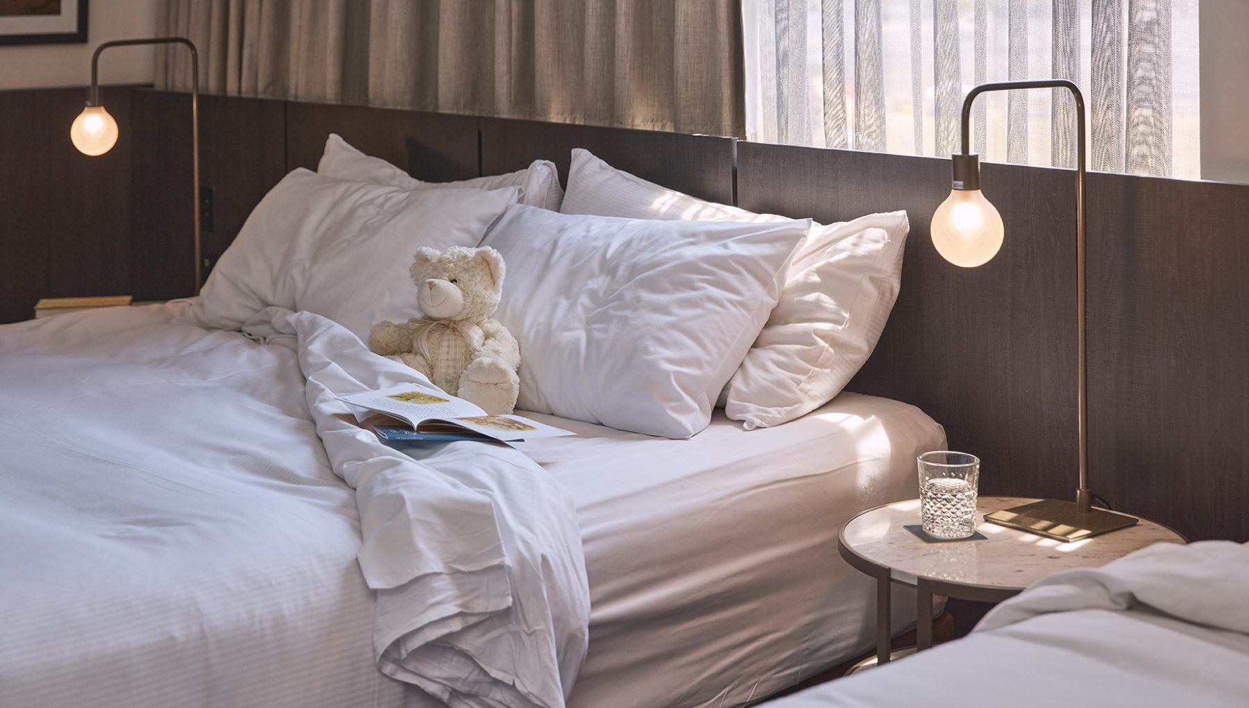A cozy bedroom scene featuring a neatly made bed with white pillows and duvet, a teddy bear, a book, and a side table with a glass.