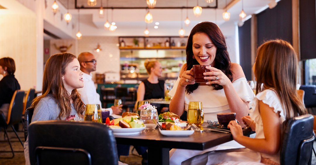 A dining scene features a woman enjoying a drink, accompanied by two girls at a table filled with various dishes and drinks.