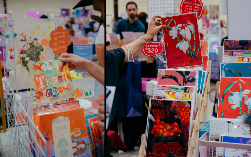 A variety of colorful art prints are displayed on wire racks at a market. A person is holding up a print while examining different options.