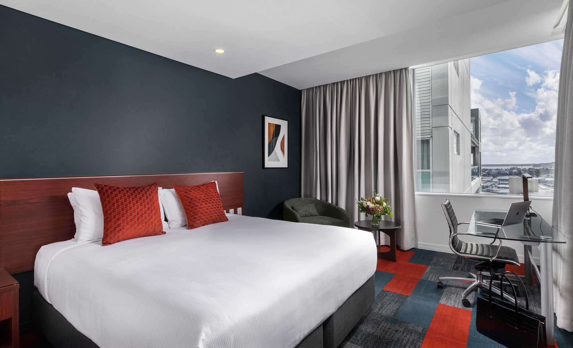 Modern hotel room with a large white bed, red cushions, a desk and laptop, and a window overlooking the city.