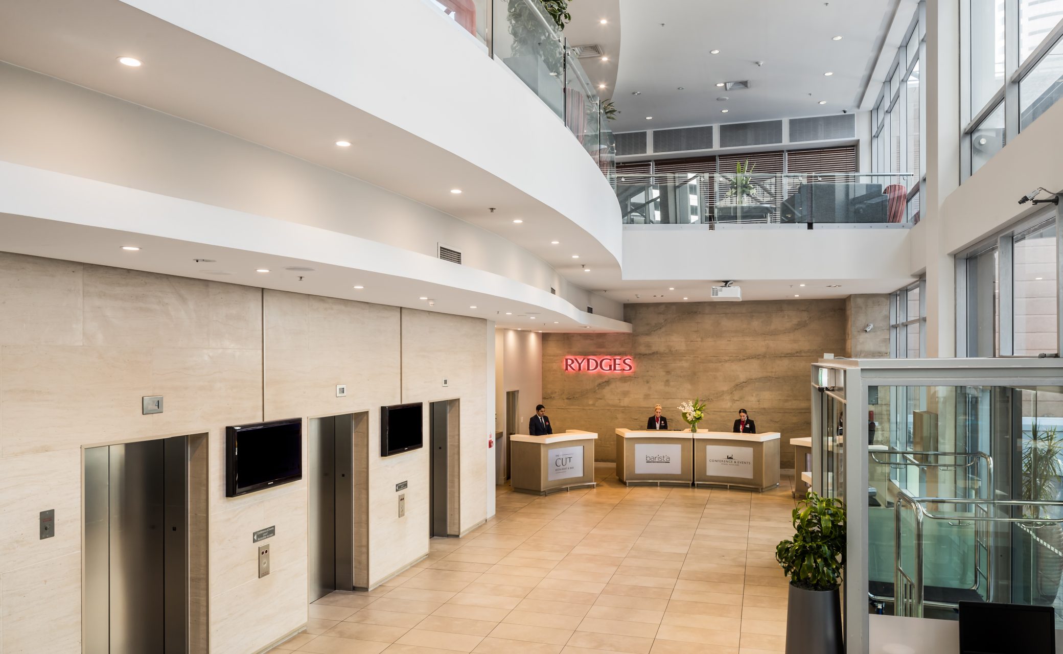 Modern hotel lobby with curved ceilings, glass elevator, tiled floors, and staff at a reception desk.