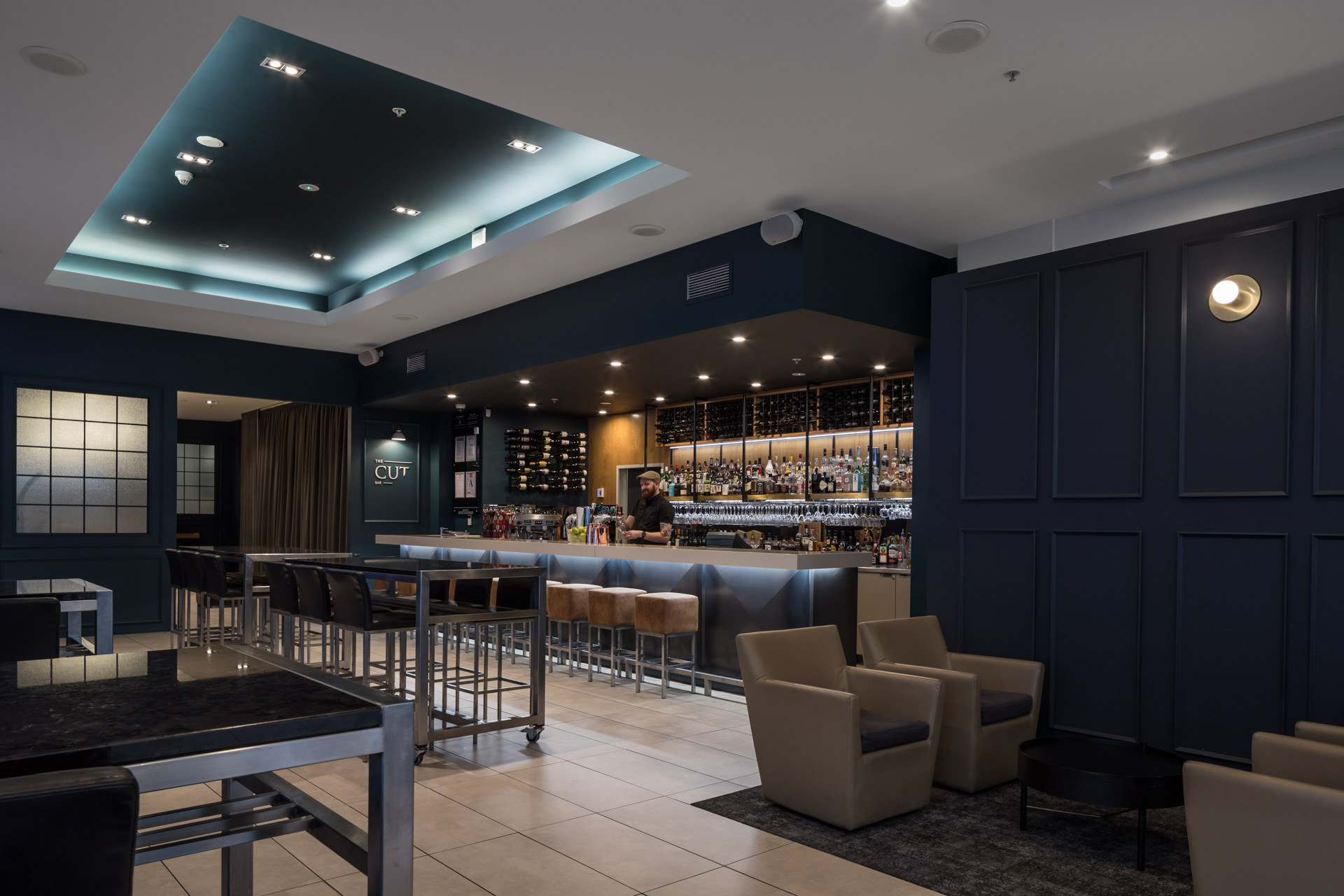 Modern dimly lit bar with a bartender behind a long counter, shelves of bottles, high tables, and lounge chairs.