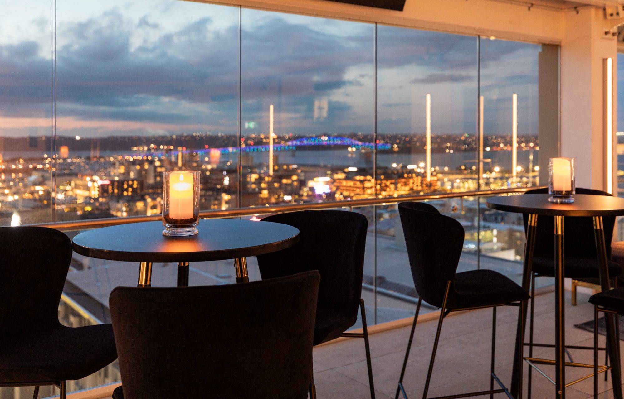 Candlelit tables and dark chairs beside floor-to-ceiling windows overlooking a city and illuminated bridge at dusk.