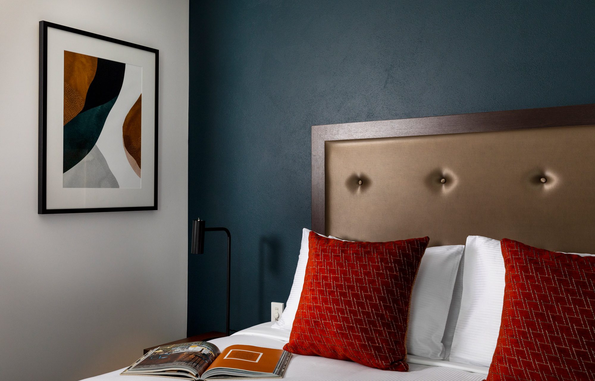 A neatly made bed with white linens and red patterned pillows sits against a teal wall, beside framed abstract art and an open book.