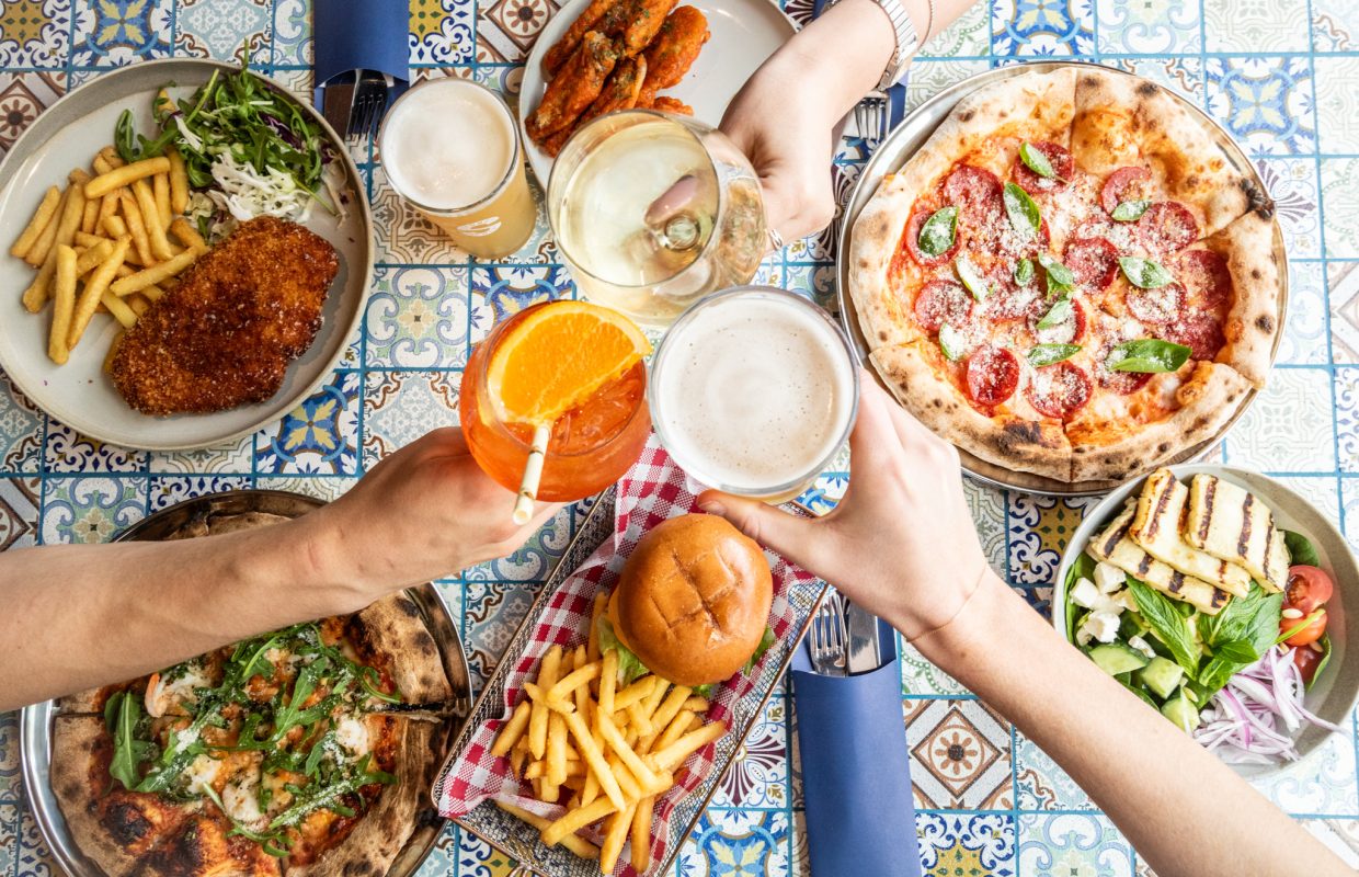 A table filled with various dishes, including pizza, fried chicken, fries, a sandwich, grilled vegetables, and drinks, surrounded by hands toasting.