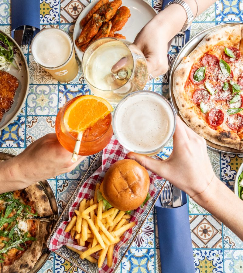 A table filled with various dishes, including pizza, fried chicken, fries, a sandwich, grilled vegetables, and drinks, surrounded by hands toasting.