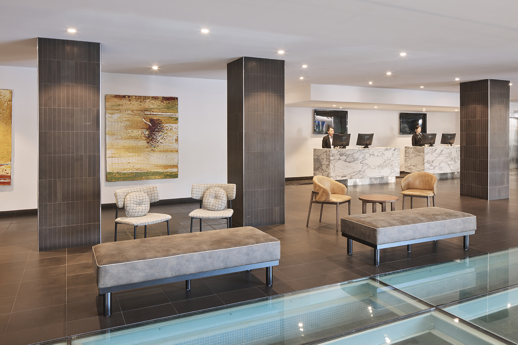 Modern lobby with marble reception desks, two staff members, lounge chairs and benches beside a glass floor feature.