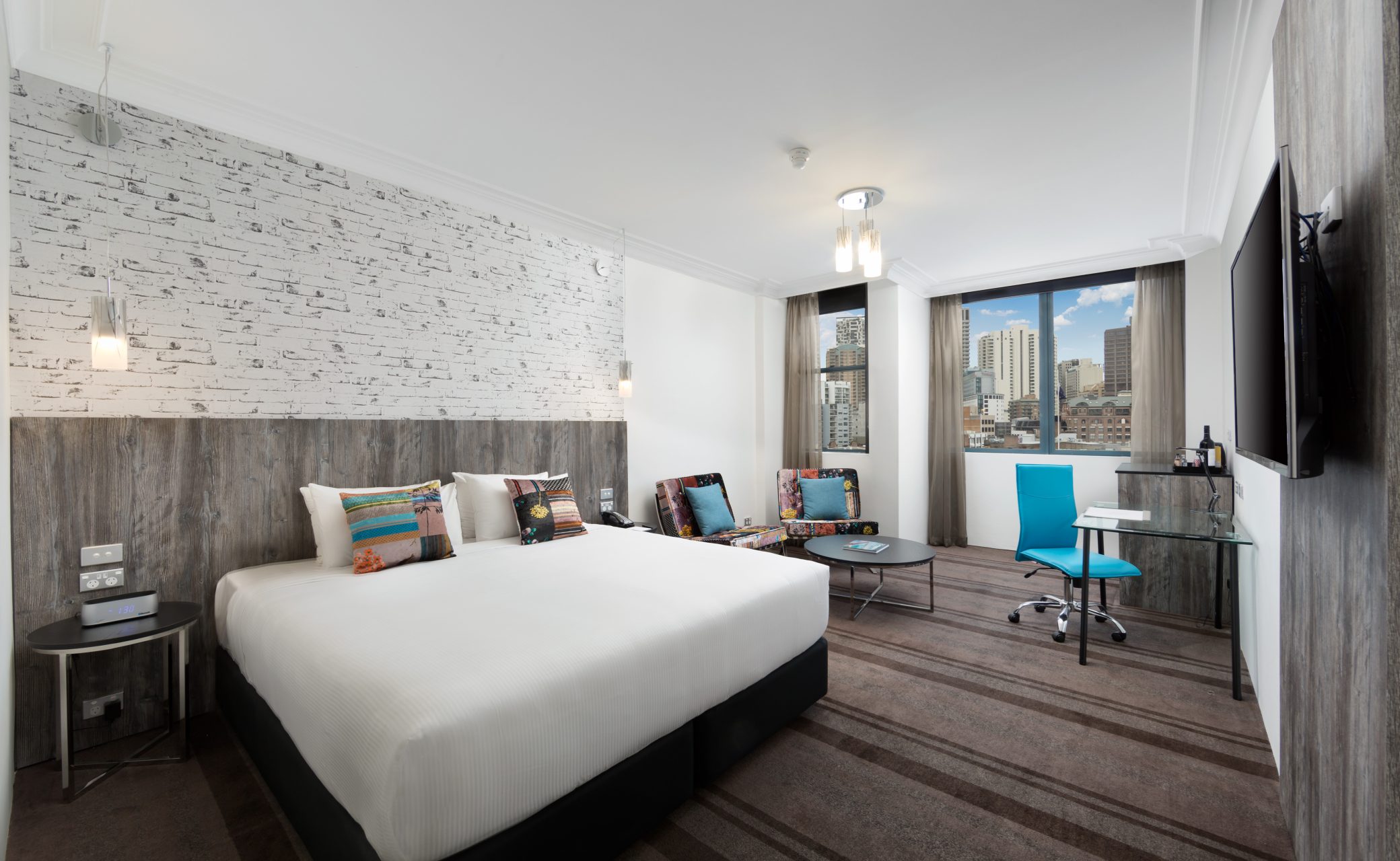 Bright hotel room with two white beds, a dark wood accent wall, city-view windows, and a turquoise lounge chair.