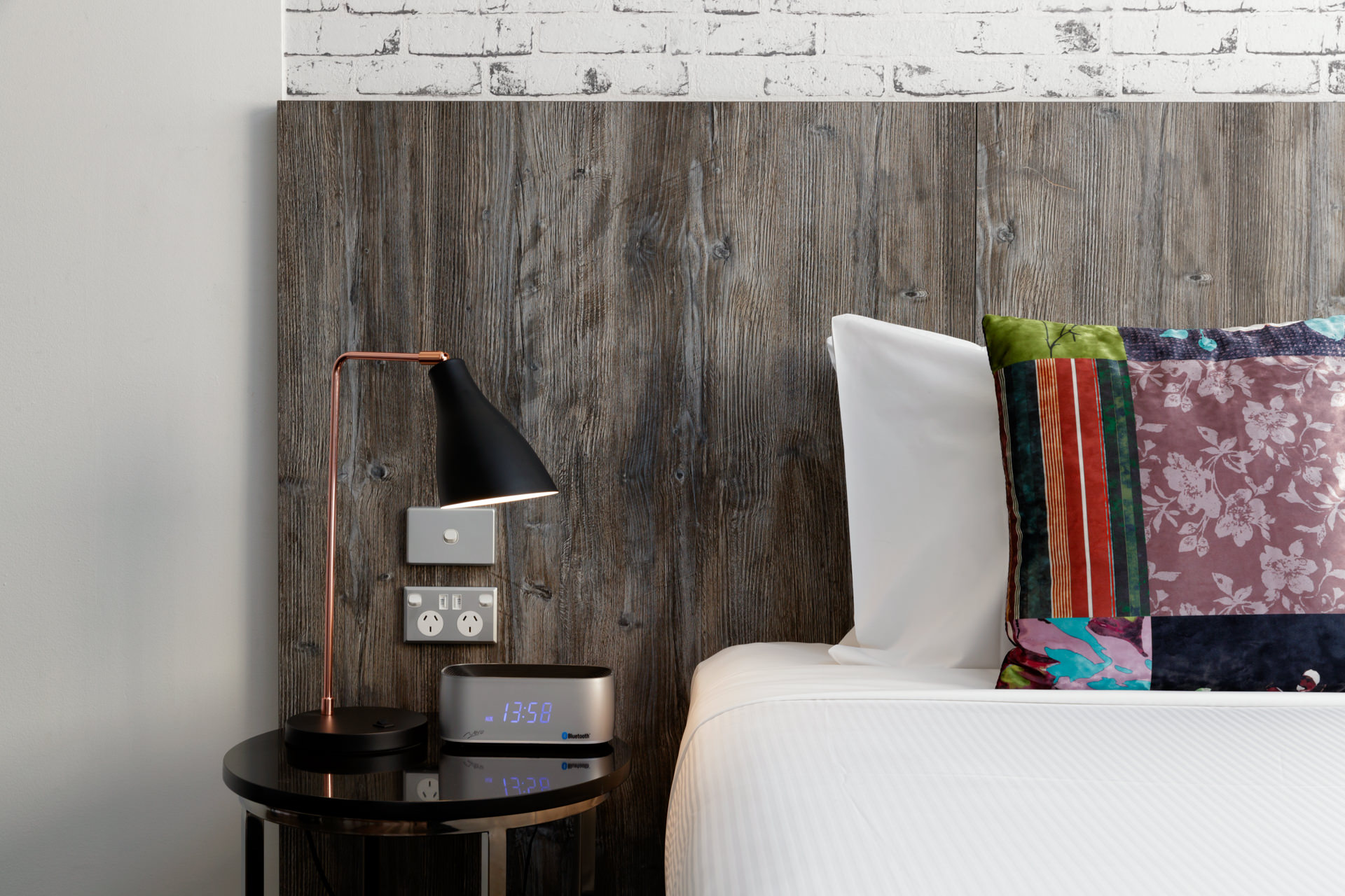A bedside table with a black-and-copper lamp and digital clock sits beside a white bed with colorful pillows.