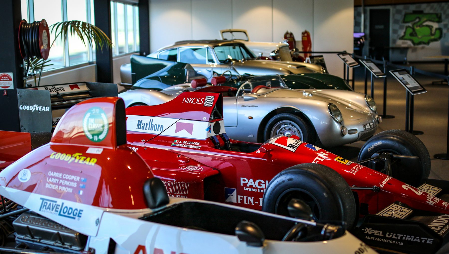 Five classic cars are displayed indoors, featuring a prominent red Formula 1 car in the foreground and a silver sports car in the background.