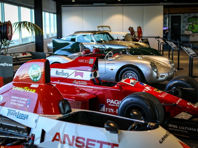 Five classic cars are displayed indoors, featuring a prominent red Formula 1 car in the foreground and a silver sports car in the background.