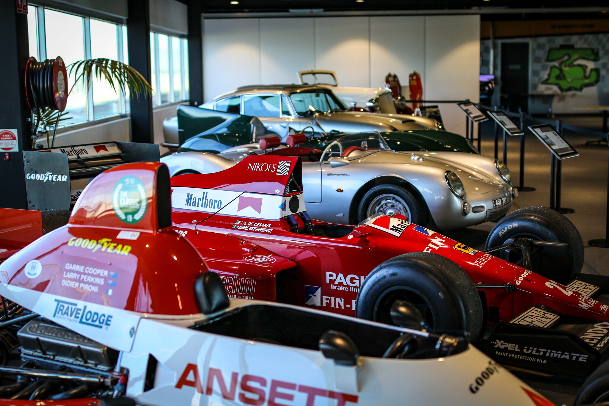Five classic cars are displayed indoors, featuring a prominent red Formula 1 car in the foreground and a silver sports car in the background.