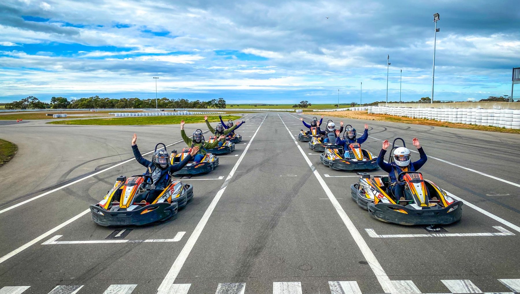 Eight go-karts are lined up on a racetrack, with drivers waving from the karts in celebration near the finish line.