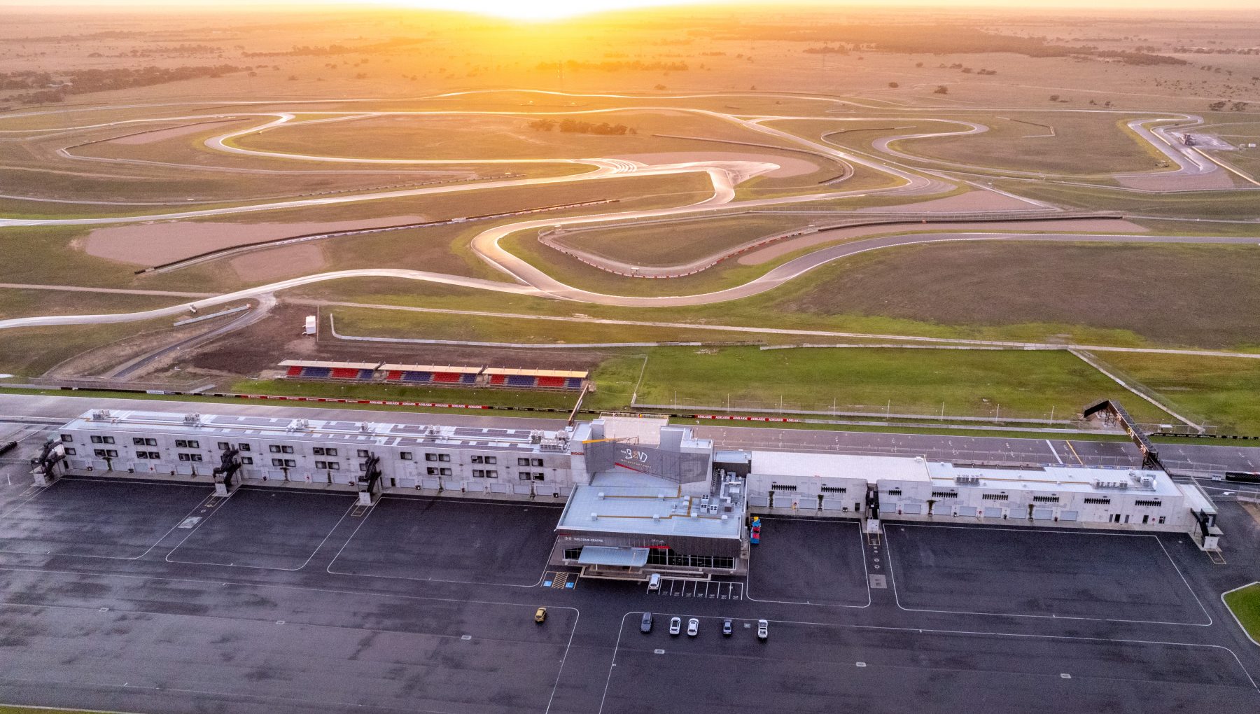 Aerial view of a racetrack with winding paths, surrounded by grassy fields and a partially visible building in the foreground.