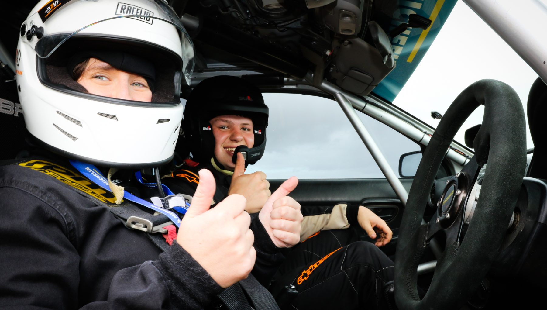 Two helmeted people sit in a rally car, smiling and giving thumbs-up beside the steering wheel.