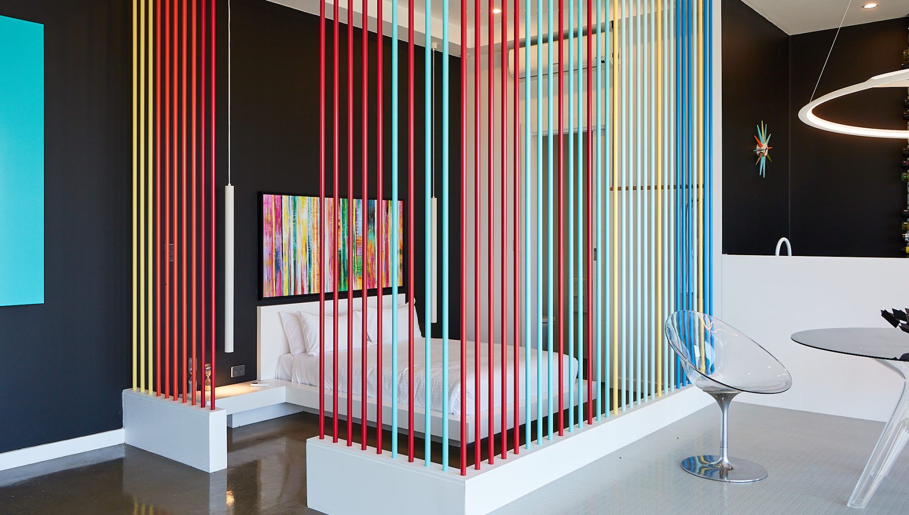 A modern bedroom features a white bed surrounded by colorful vertical strips, with a black wall and contemporary art in view.