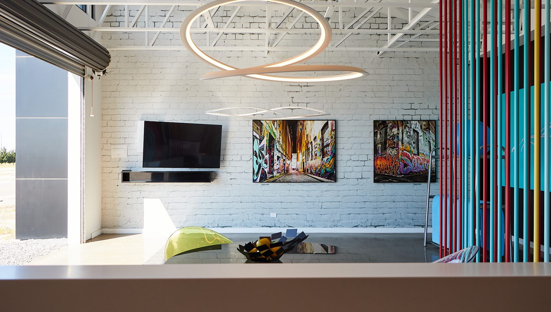 Modern interior with a white brick wall, two colorful graffiti artworks, a flat-screen TV, decorative lighting, and a unique table.