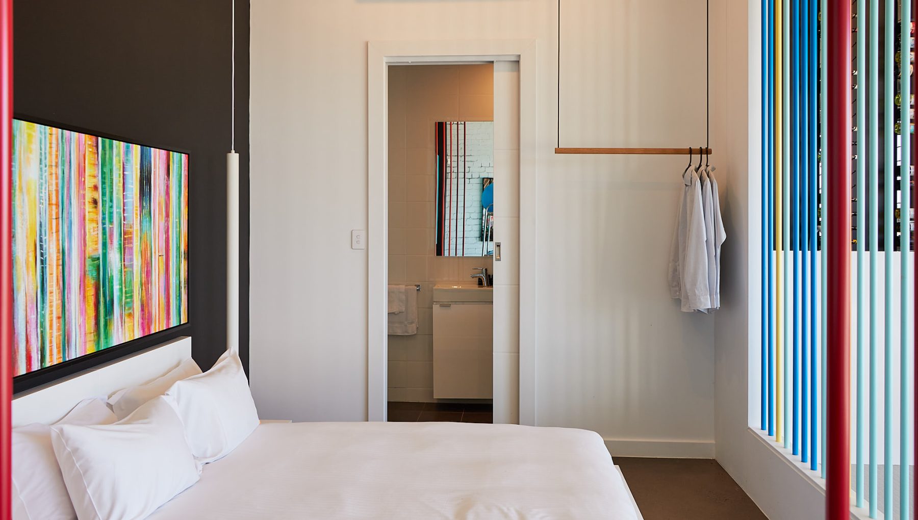 A modern, minimalist bedroom features a double bed, colorful artwork on a black wall, and a doorway leading to a bathroom. Hanging clothes and decorative window slats add a stylish touch.