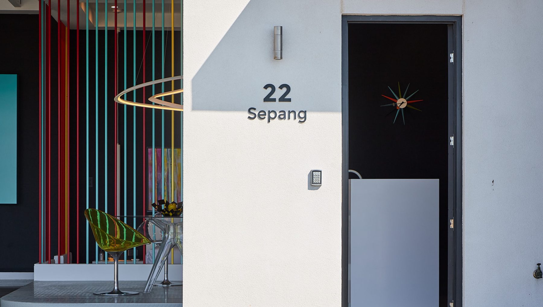 A modern entryway featuring a house number '22 Sepang', colorful vertical stripes on the wall, and a standing chair visible inside.