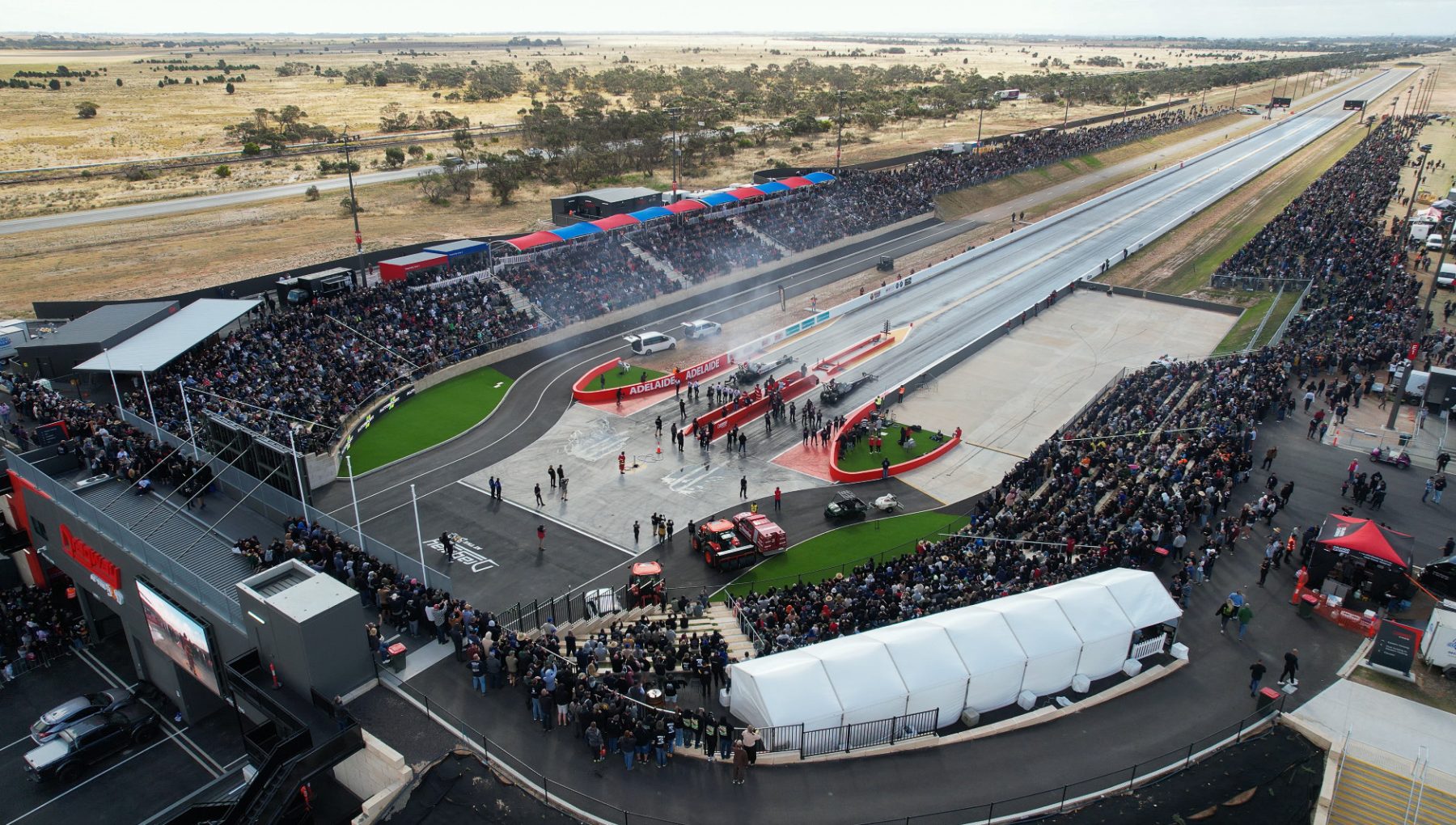 Aerial view of a drag racing track, with packed spectator stands, race cars at the start line, and smoke drifting across the strip.