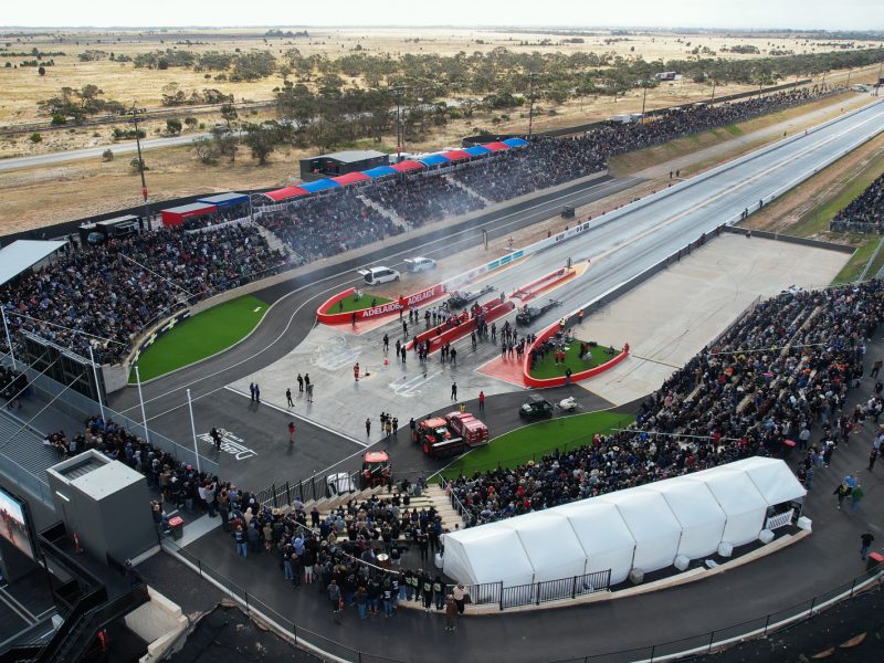 Aerial view of a drag racing track, with packed spectator stands, race cars at the start line, and smoke drifting across the strip.