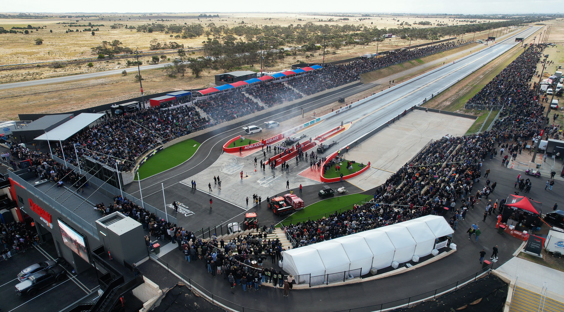 Aerial view of a drag racing track, with packed spectator stands, race cars at the start line, and smoke drifting across the strip.