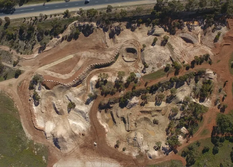 Aerial view of a large earthworks site with winding dirt tracks, excavated ground, scattered trees, and a road along the top edge.