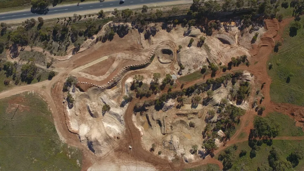 Aerial view of a large earthworks site with winding dirt tracks, excavated ground, scattered trees, and a road along the top edge.