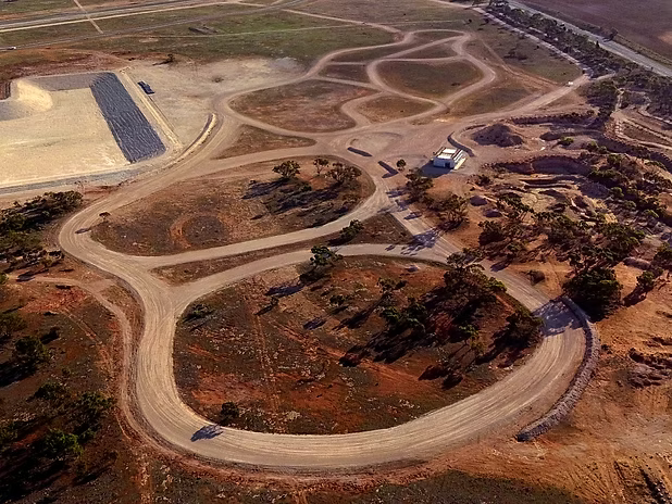 Aerial view of winding dirt roads looping through dry terrain, with scattered trees, a small building, and a paved rectangle.