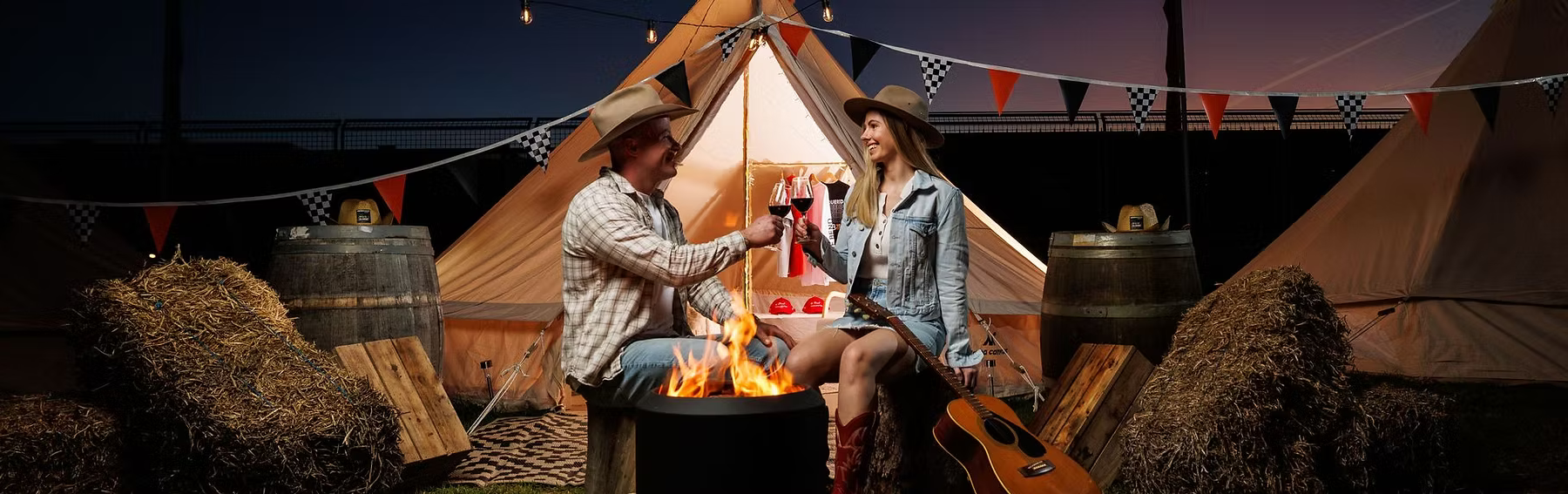 Two people clink wine glasses beside a campfire in front of a tent, with a guitar, hay bales, and bunting nearby.