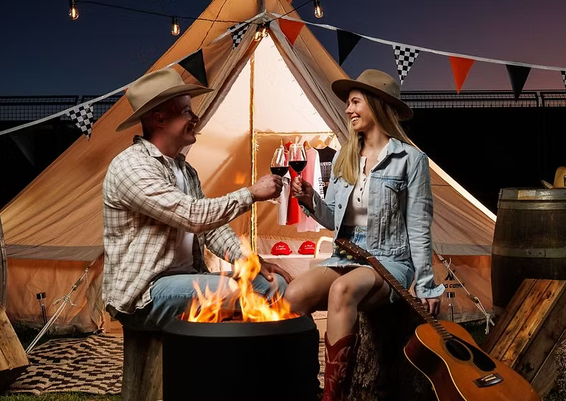 Two people clink wine glasses beside a campfire in front of a tent, with a guitar, hay bales, and bunting nearby.