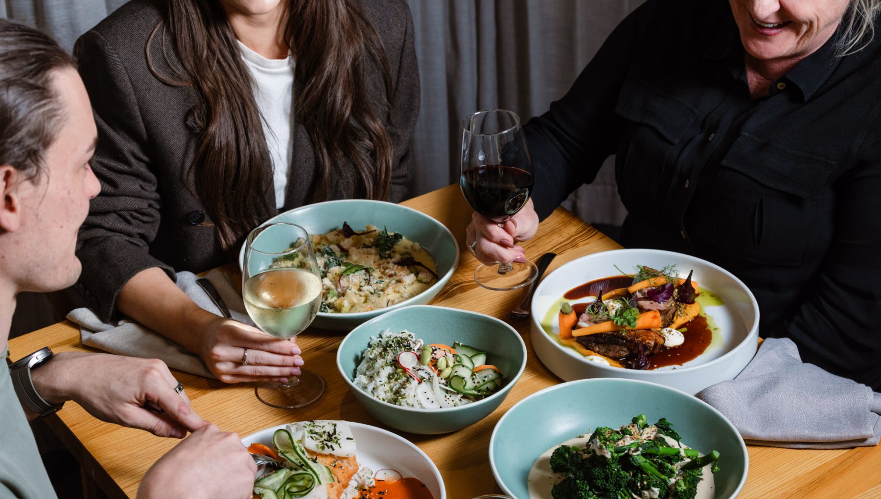 Three diners share plated meals and wine at a wooden restaurant table.