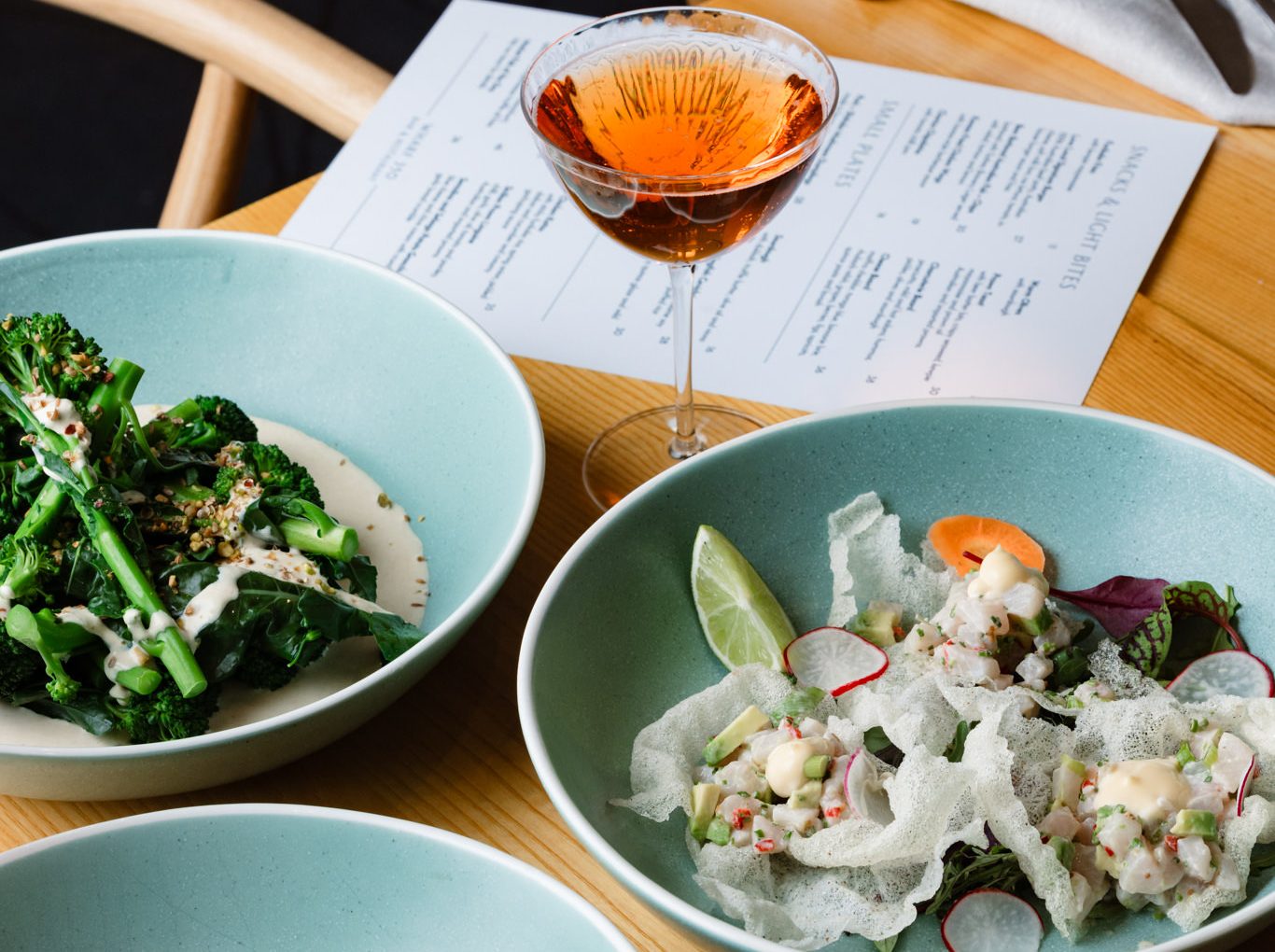 Three turquoise plates of greens, ceviche, and flatbread sit on a wooden table beside an amber drink and menu.