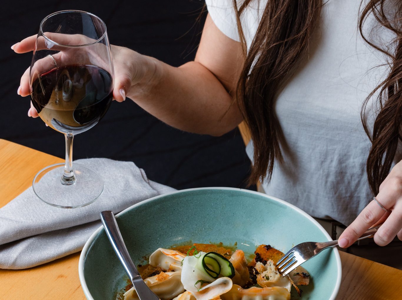 Woman holding a glass of red wine while eating dumplings at a restaurant table.