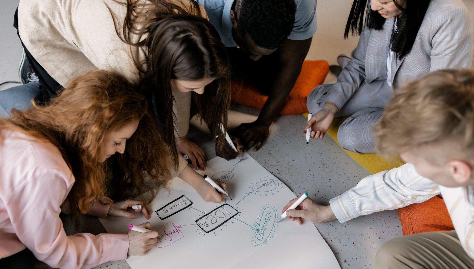 A group of children gathers around a table, drawing and writing on large sheets of paper with markers.