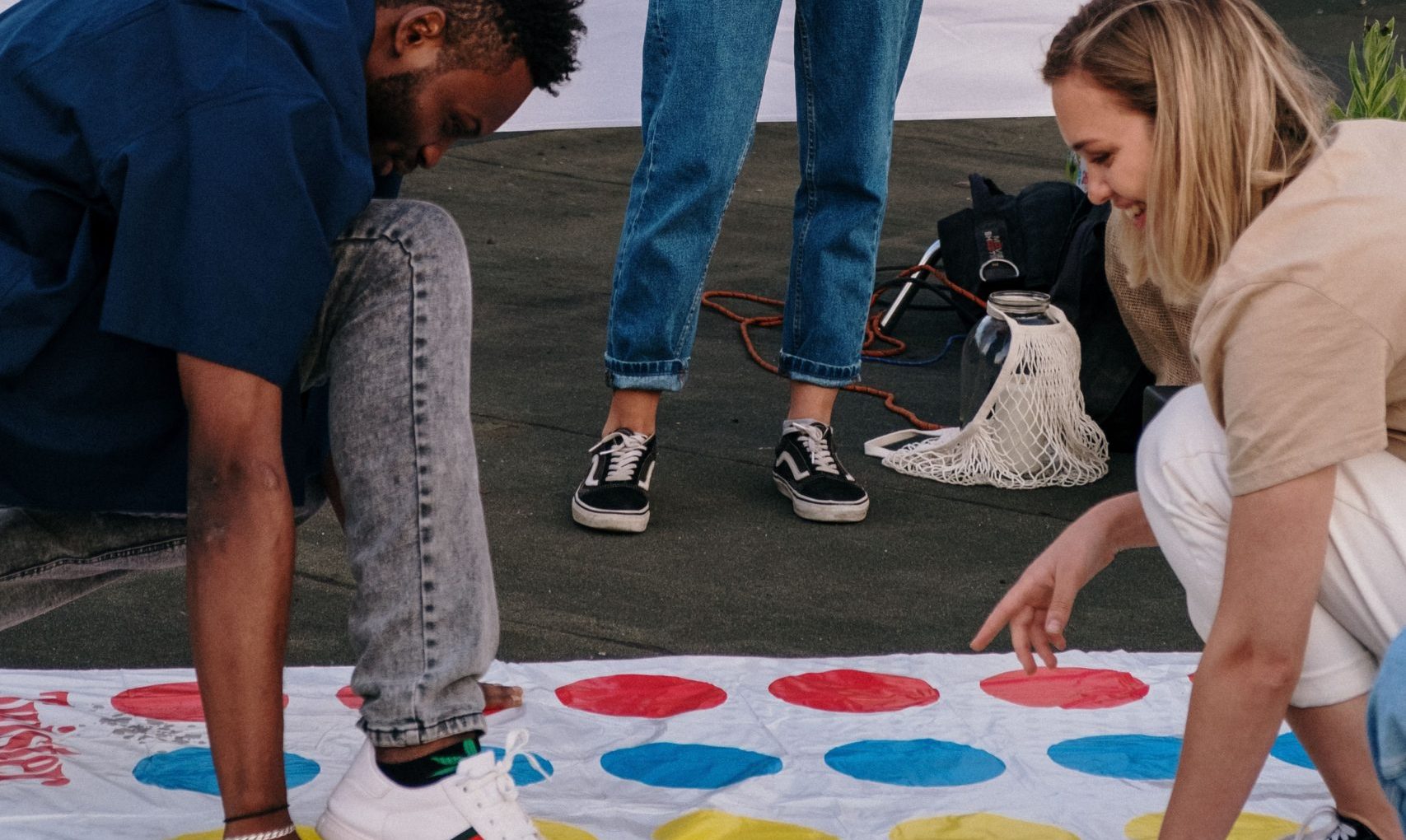 A smiling woman holds a game spinner while friends crouch on a colorful Twister mat beneath hanging lights.
