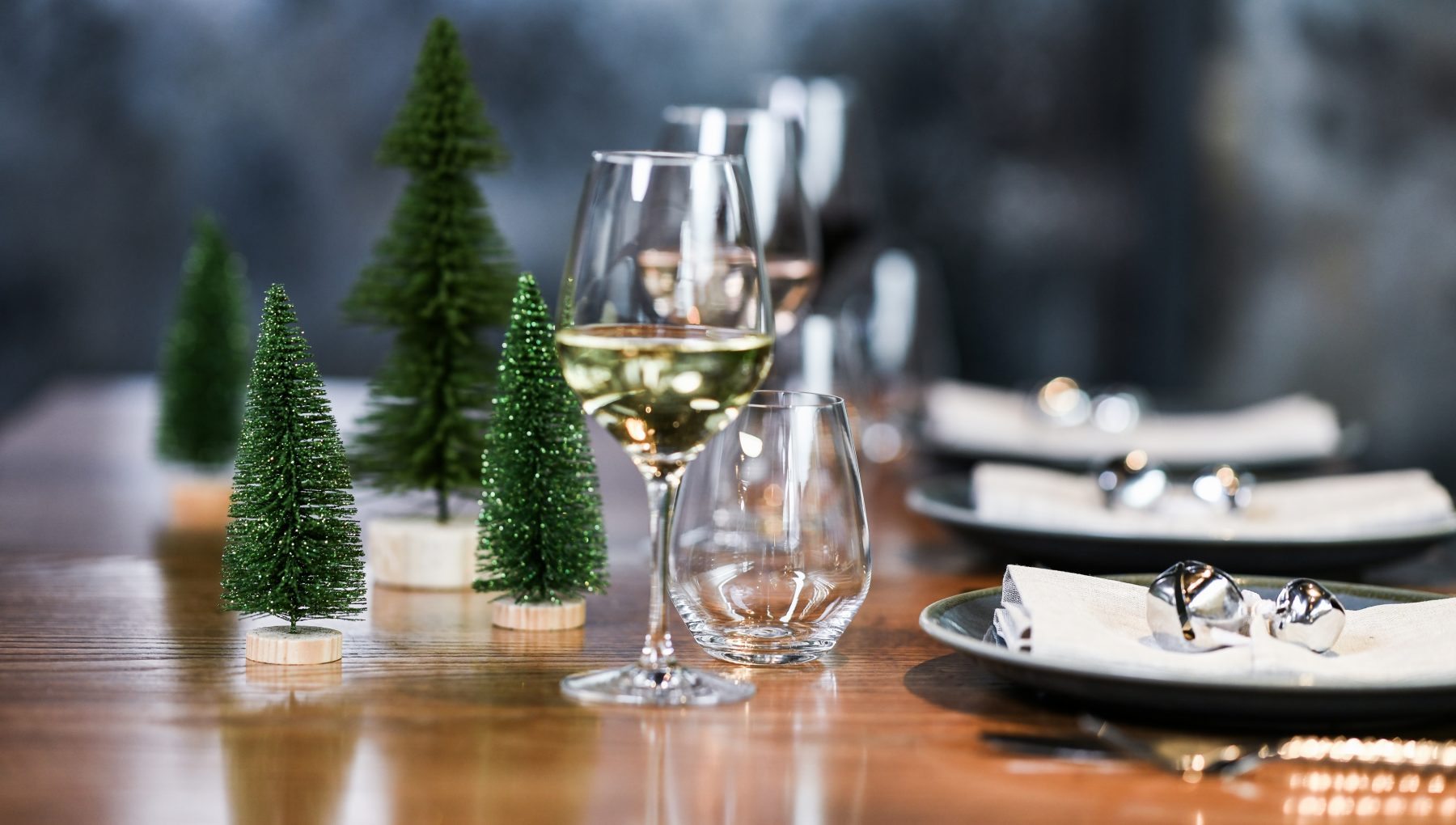 Decoratively set dining table with small green trees, wine glasses, and plates with napkins and silver bells.