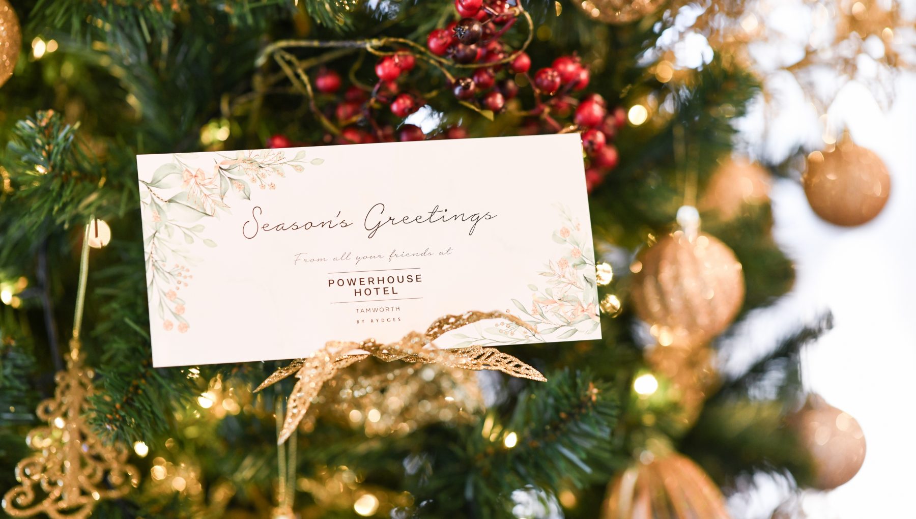 A floral-bordered Season’s Greetings card rests among lit evergreen branches, red berries, and gold ornaments.