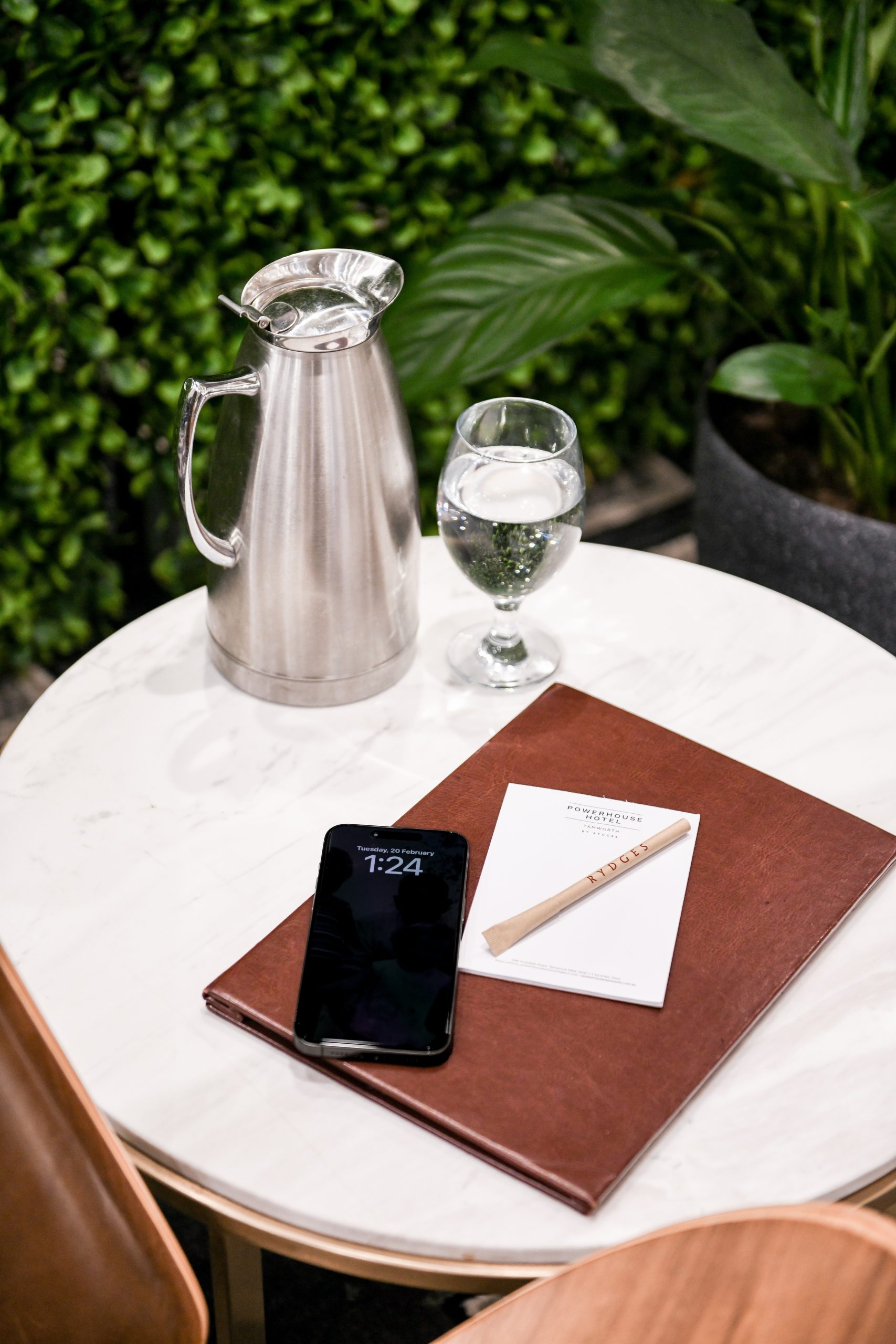 A silver pitcher and a glass of water sit on a marble table next to a smartphone and a brown menu with a notepad. Green foliage is in the background.