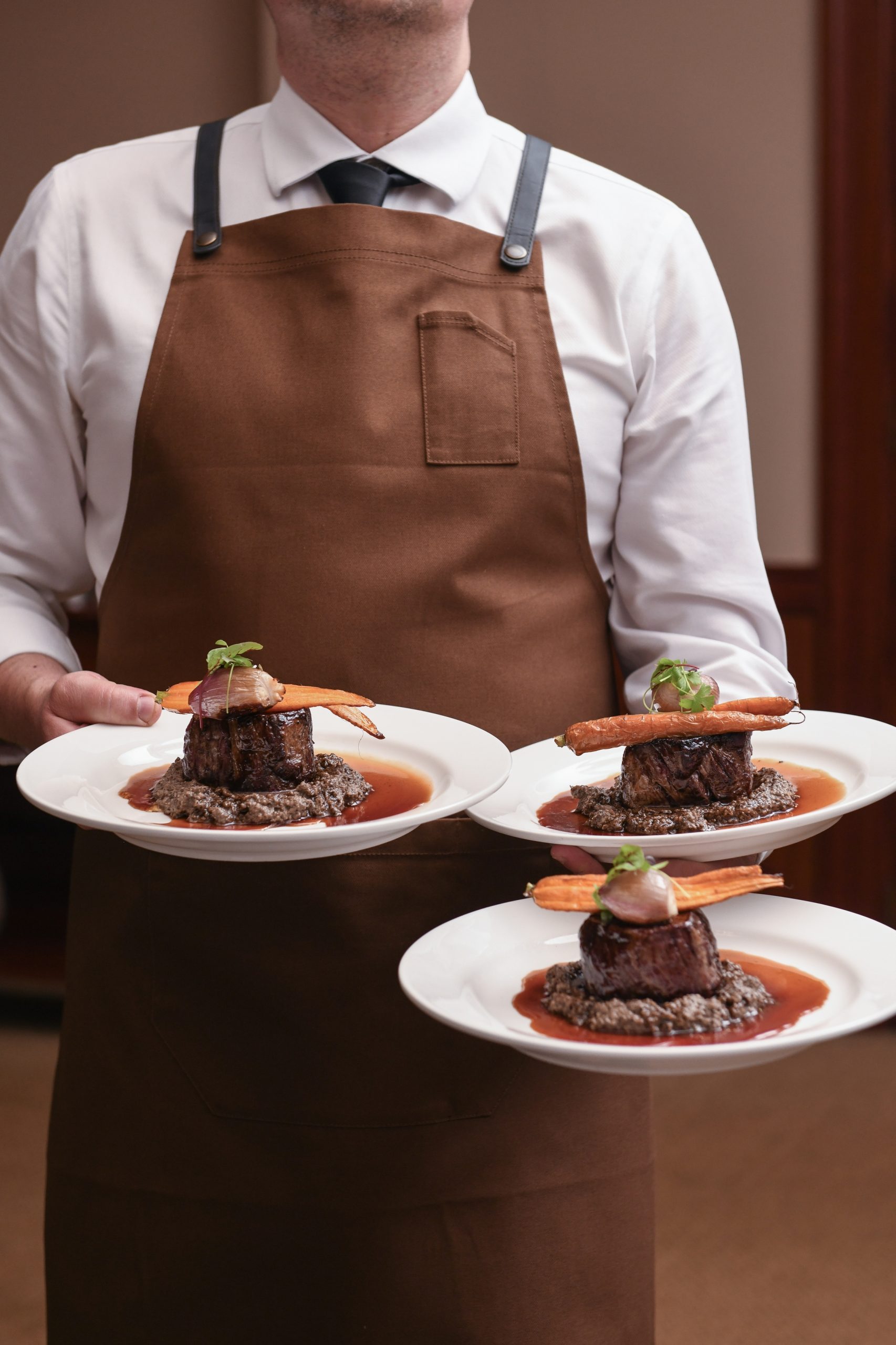 A waiter in a brown apron holds three plates, each featuring a piece of meat on a bed of sauce with carrots and garnish.