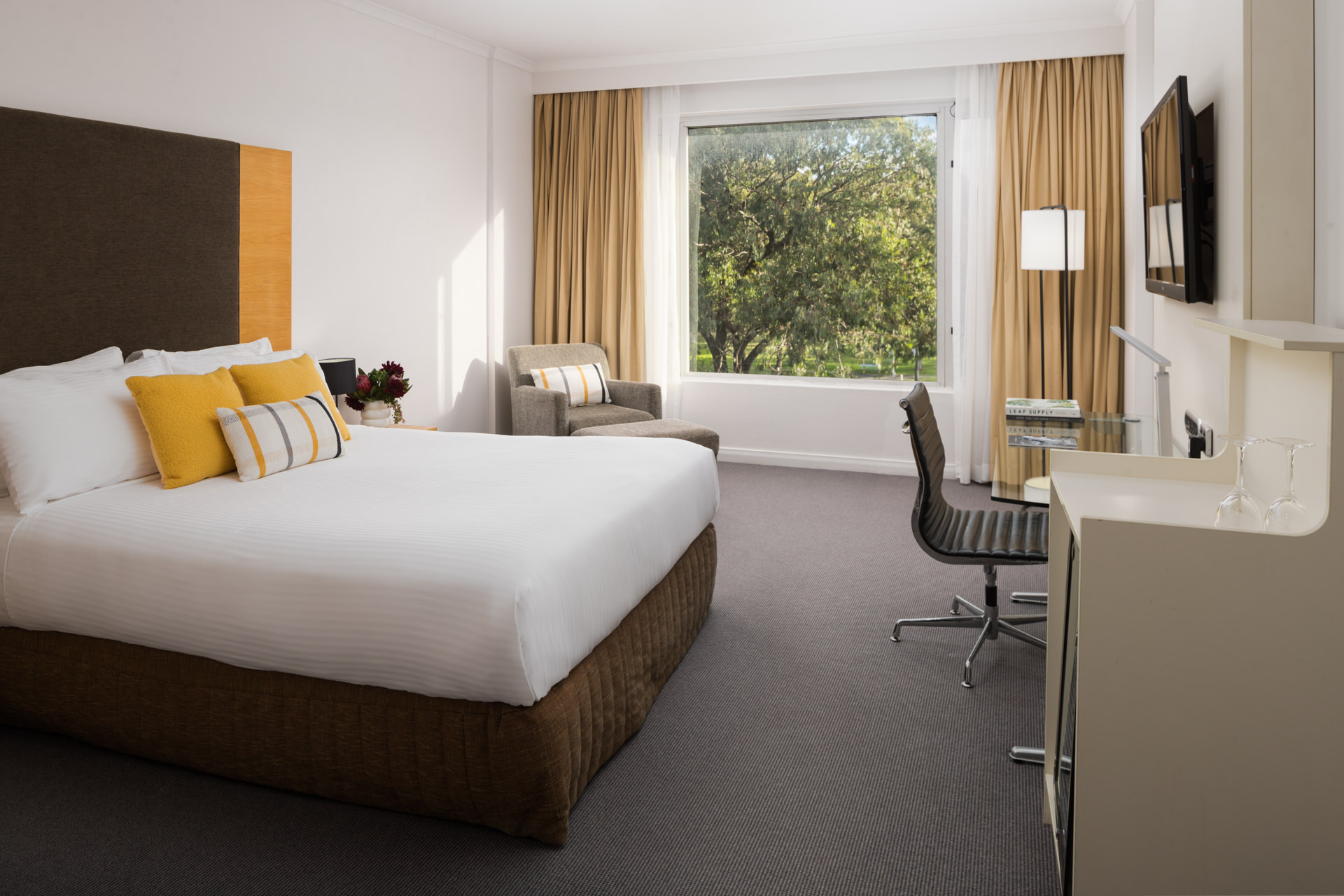 A spacious hotel room featuring a queen-sized bed, armchair, desk with a chair, and large window with outdoor views.