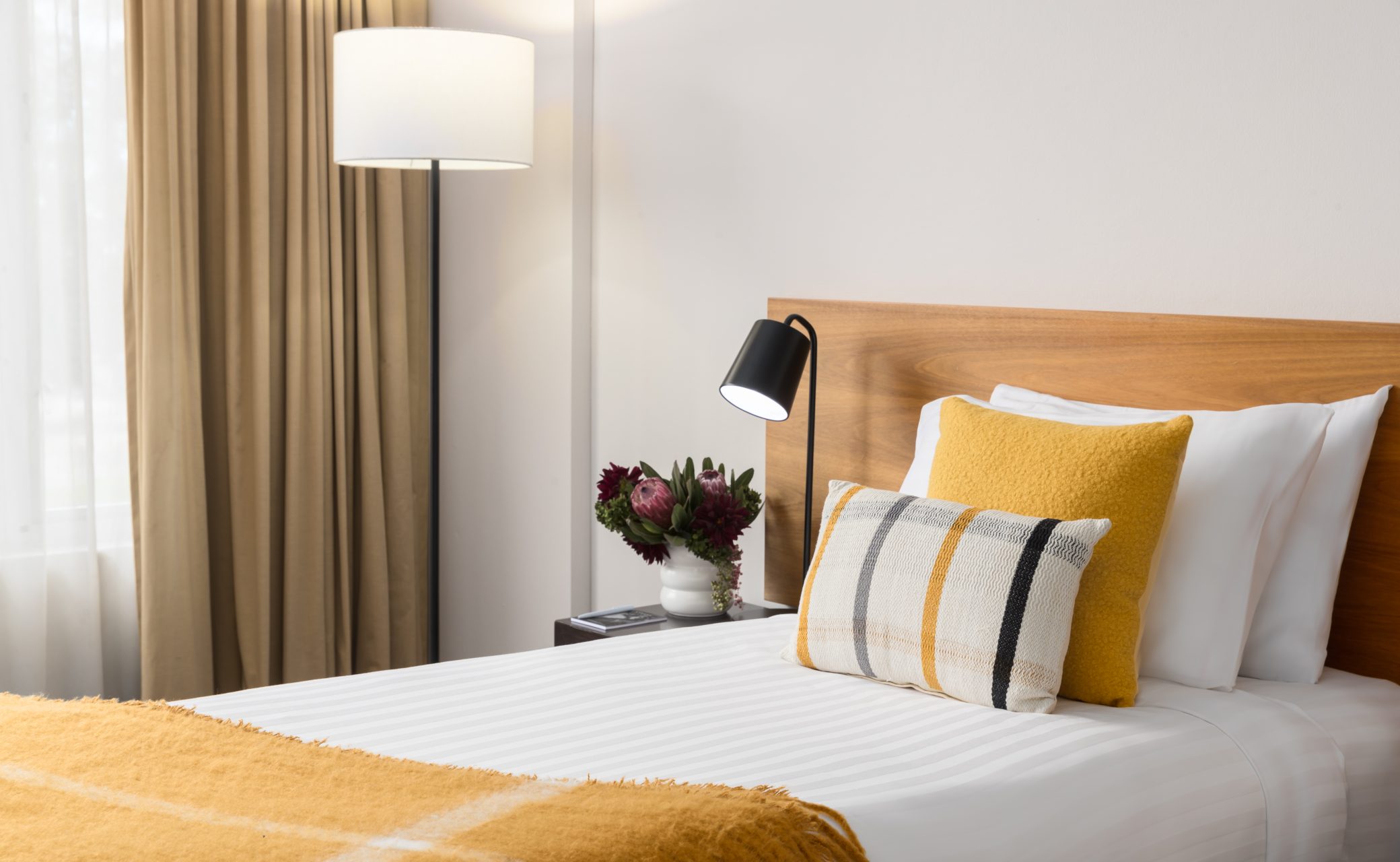 A cozy bedroom featuring a bed with white linens, yellow and patterned pillows, and a decorative throw. A floor lamp and a vase of flowers are beside the bed.